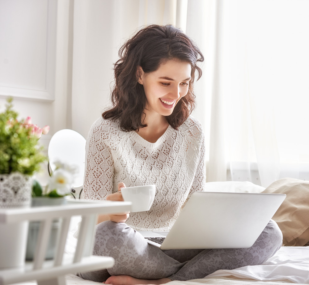 This is an image of a smiling young woman sitting on a bed in a sunlit room using a laptop computer.